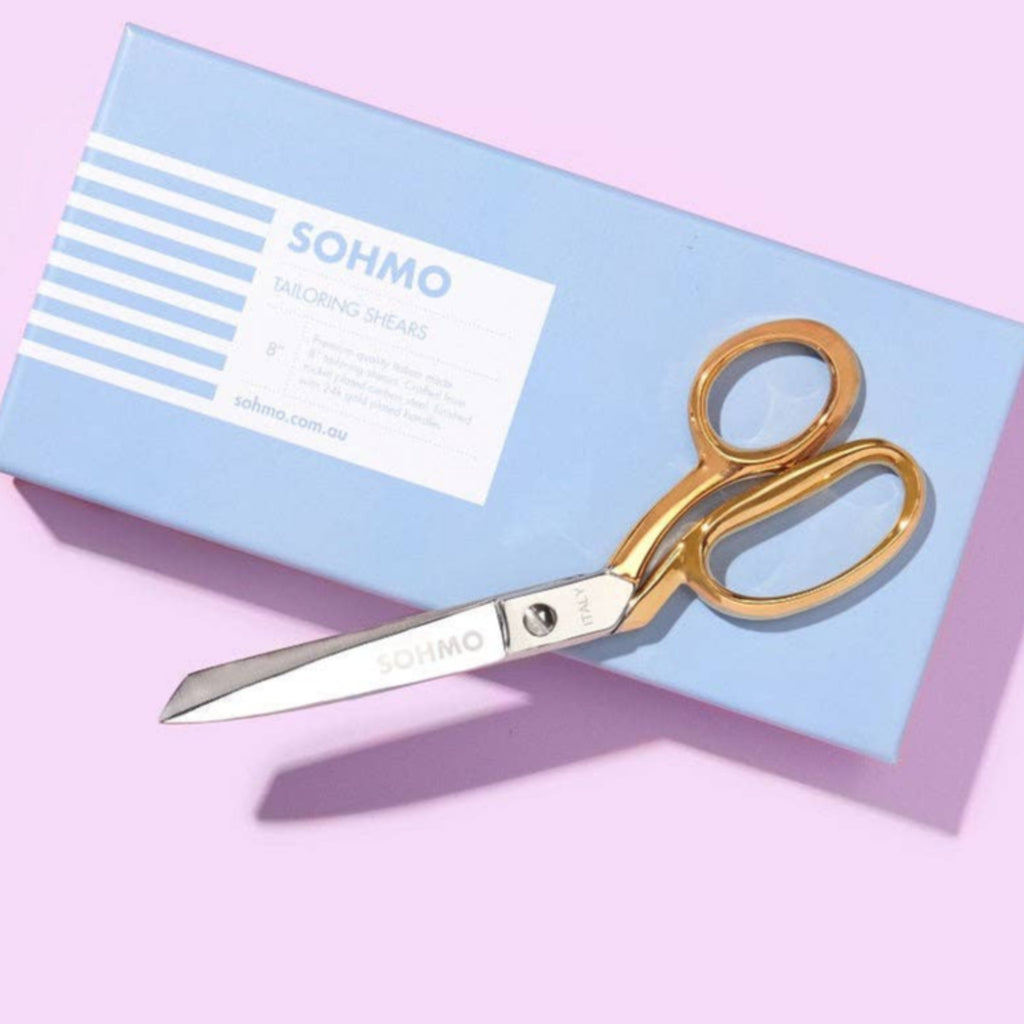 Gold scissors on a blue box with 'Sohmo' branding against a pink background