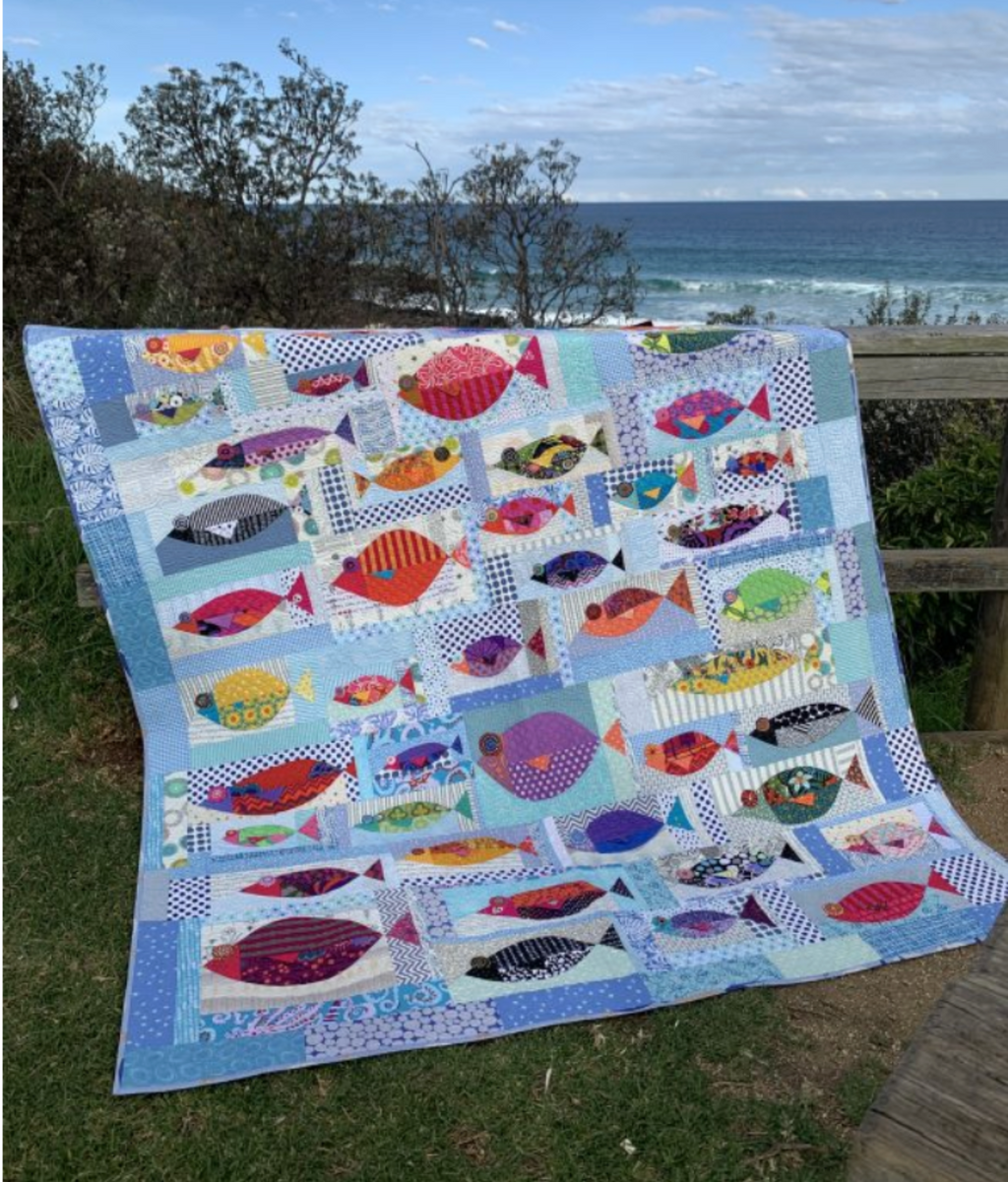 Colorful fish-patterned quilt on a grassy area with ocean view