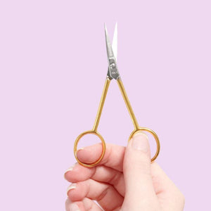 Hand holding a pair of gold-handled scissors against a light purple background