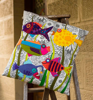 Decorative pillow with colorful fish design on a wooden stand against a stone wall.