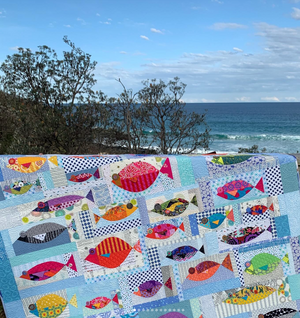 Colorful fish-patterned quilt on a bench with a beach and ocean view.