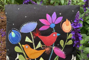 Decorative pillow with colorful embroidered birds and flowers on a black background, surrounded by greenery.